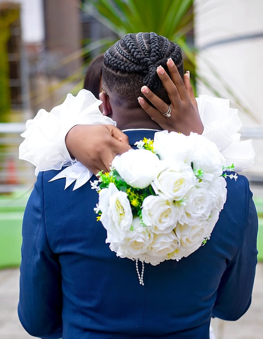 Bride embracing a groom in a blue suit, holding white roses